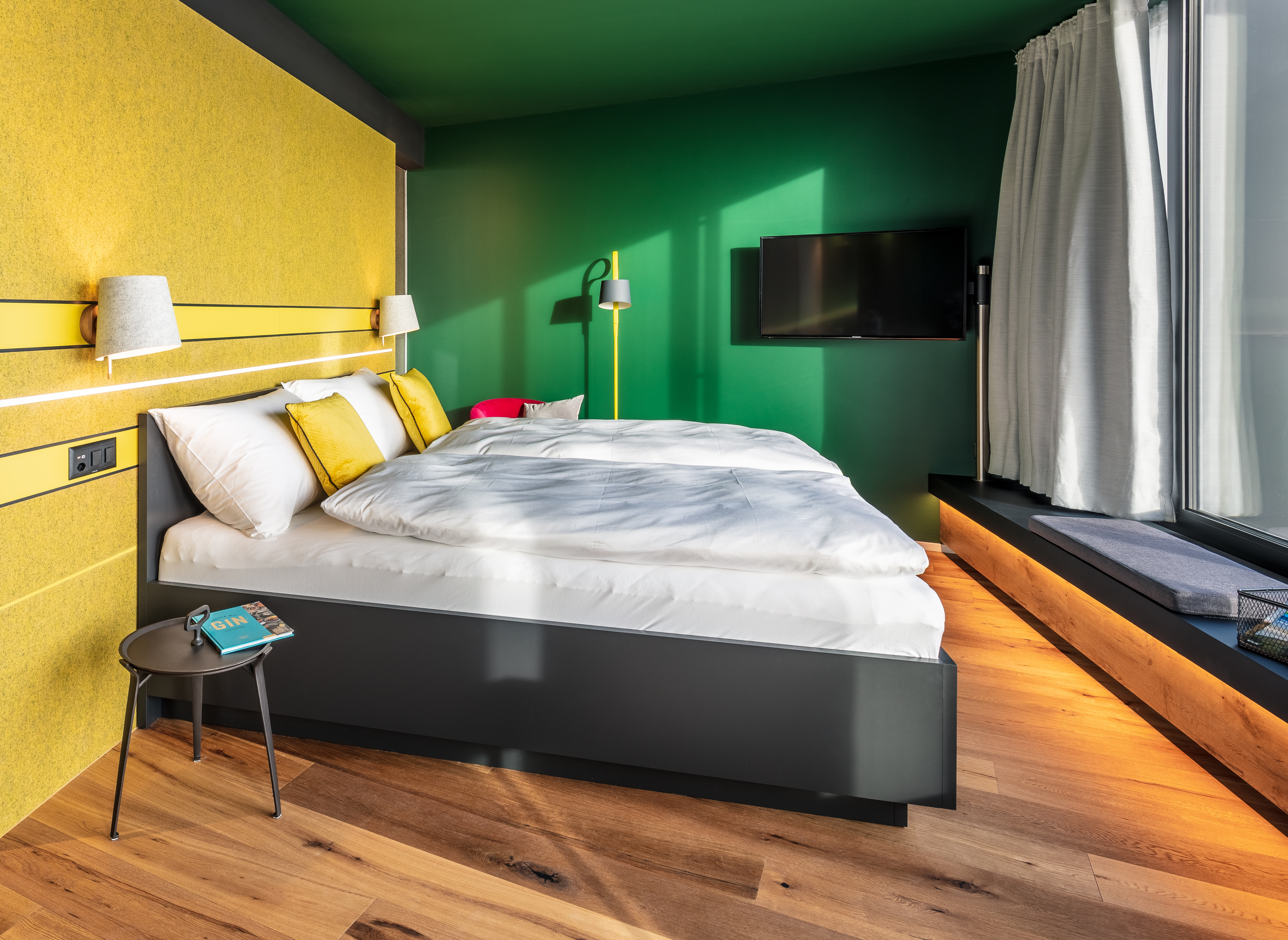 Zimmeransicht mit Doppelbett, das mit weißer Bettwäsche und gelben Kissen bezogen ist. Auffallend die gelbe Wand hinter dem Bett und eine grüne Wand mit Fernseher. Ein kleiner Tisch mit Buch steht neben dem Bett. Helles Holzparkett rundet das Bild ab.